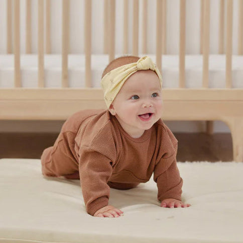 Baby in a brown outfit and yellow headband crawling on a beige mat in front of a wooden crib.