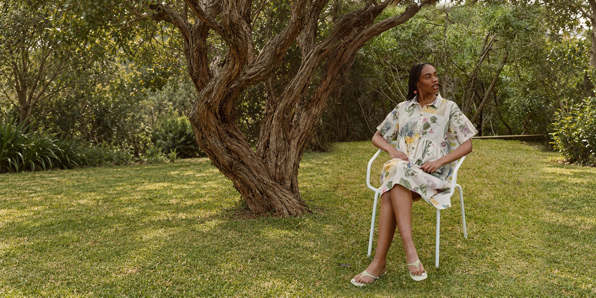 Model wearing a cream botanical-print shirt dress, seated beneath a garden tree.